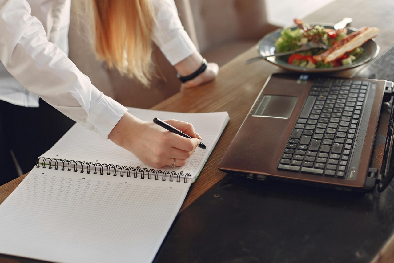 Woman writing in a food diary notebook next to a laptop and meal
